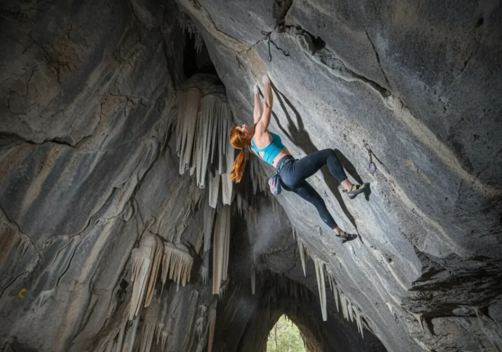 A female climber with a spectacular physique performs a difficult move on a massive cave ceiling as other climbers watch from below.
