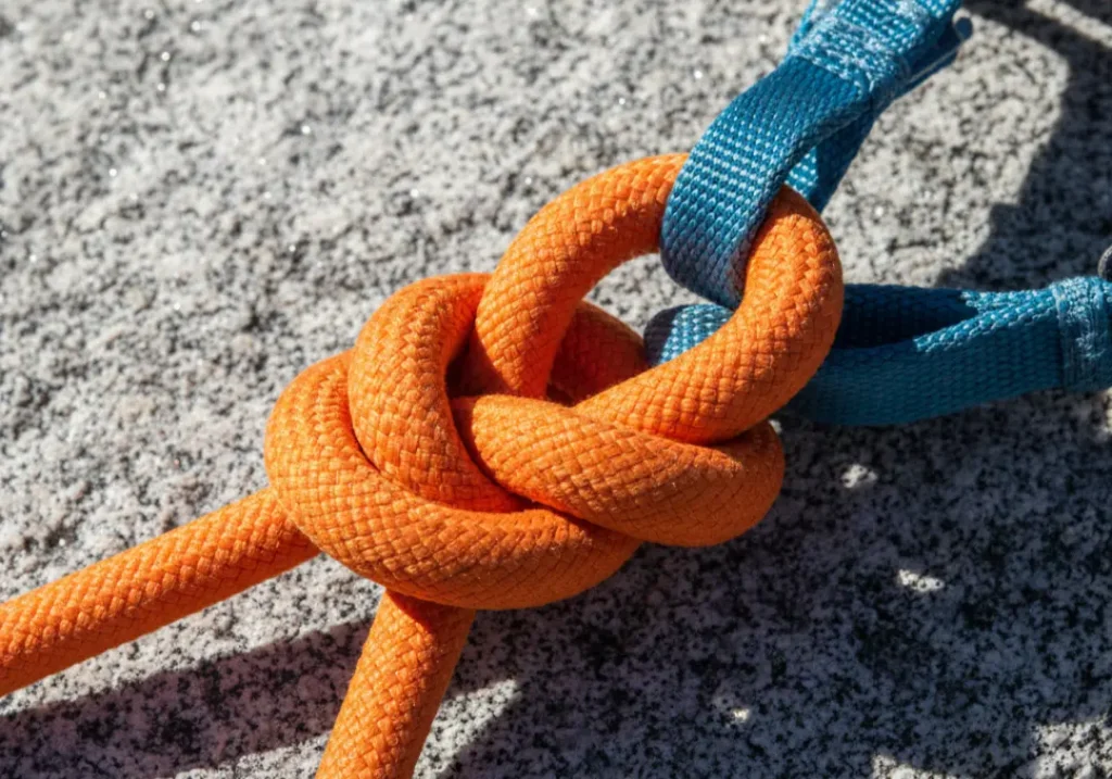 A close-up of a perfectly tied orange climbing rope using a figure-eight knot connected to a blue climbing harness, resting on a rock.