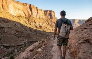 The Access Fund: A Climber’s Guide to Stewardship A climber with a backpack hiking on a dusty trail towards a large sandstone cliff face during sunset.
