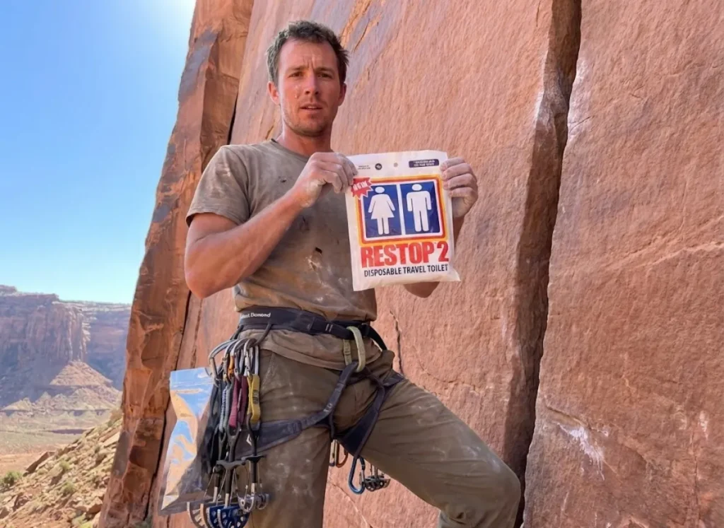 A climber at Indian Creek holding a Restop 2 waste bag against a backdrop of red sandstone cliffs and blue sky.