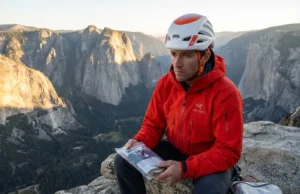 Mastering How to Poop Outdoors From Crag to Big Wall A professional climber standing on a high alpine ledge at sunset, holding a waste management kit, wearing a red Arc'teryx jacket and Petzl helmet, overlooking a mountain range.