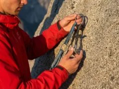 Resling Cams Safely: The Definitive Repair vs Retire Guide Professional climber wearing an Arc'teryx jacket inspecting a Black Diamond Camalot at the base of a granite cliff during golden hour.