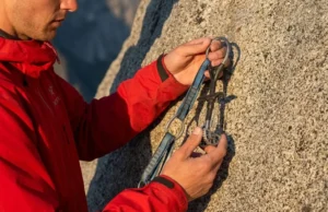 Resling Cams Safely: The Definitive Repair vs Retire Guide Professional climber wearing an Arc'teryx jacket inspecting a Black Diamond Camalot at the base of a granite cliff during golden hour.