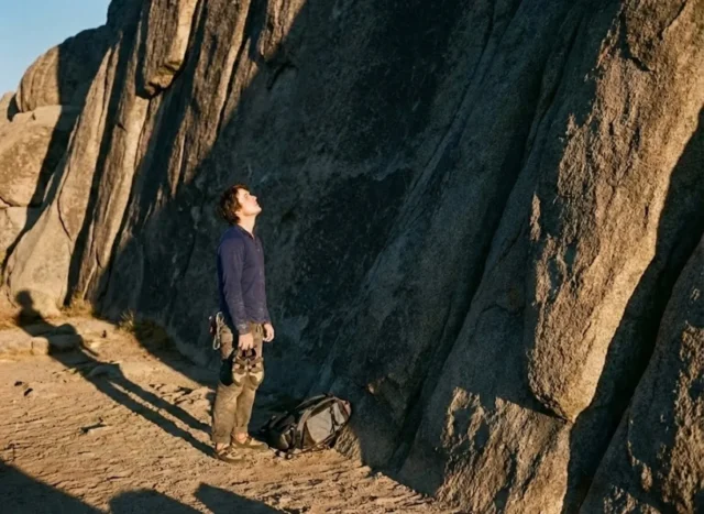 sociology-of-climbing A climber stands at the base of a large granite cliff at sunset, preparing gear for a route.