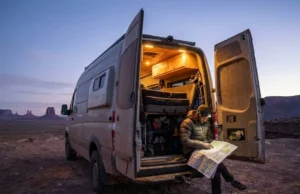 Van Life for Climbers: Engineering a Mobile Basecamp A dusty 4x4 adventure van parked on a rocky desert cliff at dusk with rear doors open, illuminating a climber reading a map on the bumper.