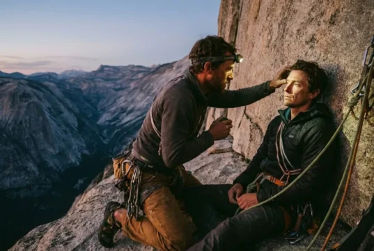 Wilderness First Aid for Climbers: Vertical Field Guide A climber on a high rock ledge performs a medical assessment on their partner during sunset.