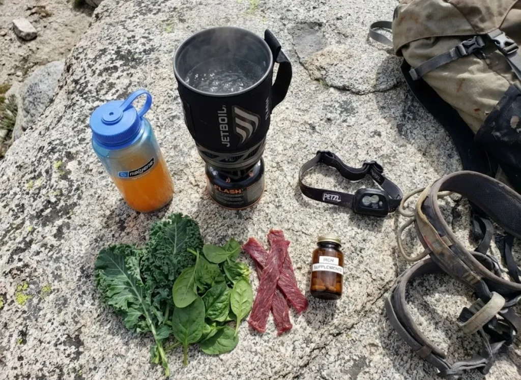 A basecamp meal setup on a rock featuring a Jetboil stove, Nalgene bottle, and iron supplements.