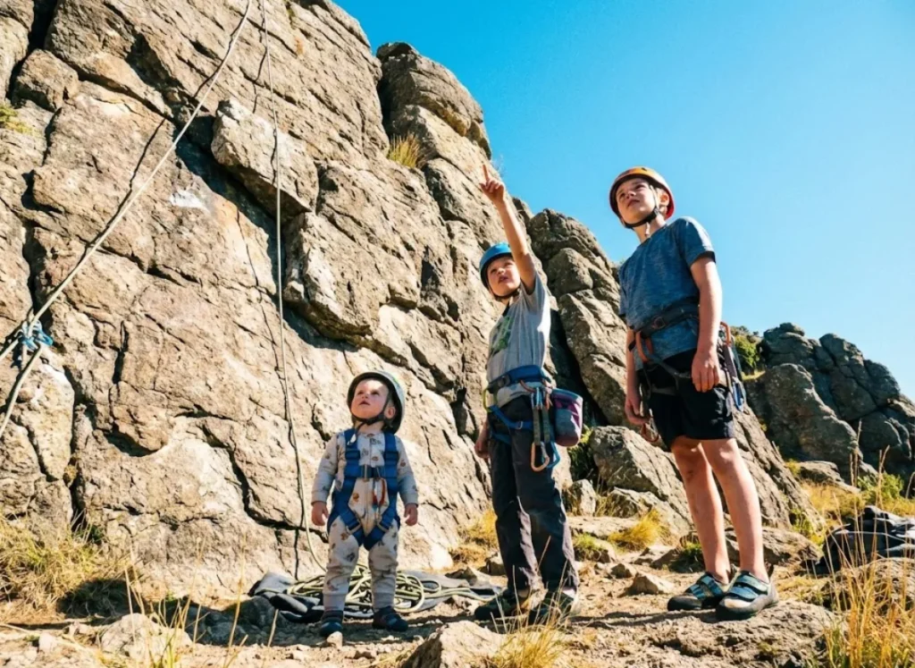 Three children of different ages wearing climbing gear standing at an outdoor crag.