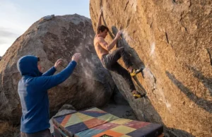 Bouldering Grades Explained: The V0-V17 Benchmarks Climber wearing La Sportiva Solution shoes scaling a highball boulder in Bishop with a spotter in a Patagonia fleece and Organic Climbing crash pads below.