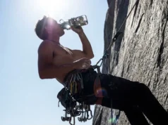 Hydration for Climbers: Dosage, Gear & DIY Recipes A rock climber at a high exposure hanging belay drinking water from a durable bottle amidst climbing gear.
