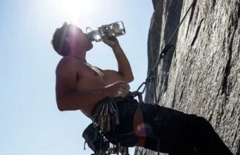 Hydration for Climbers: Dosage, Gear & DIY Recipes A rock climber at a high exposure hanging belay drinking water from a durable bottle amidst climbing gear.