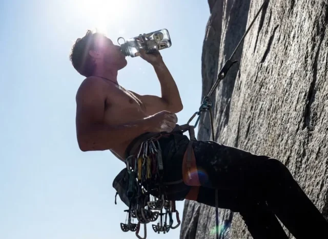 hydration-climbers-dosage-gear A rock climber at a high exposure hanging belay drinking water from a durable bottle amidst climbing gear.