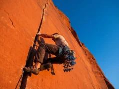 Indian Creek Climbing Guide: Rack, Skills & Rules A climber ascending a vertical red sandstone crack at Indian Creek, Utah, wearing tape gloves and carrying a heavy rack of camming devices during sunset.