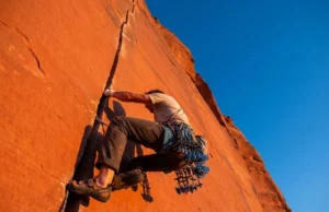 Indian Creek Climbing Guide: Rack, Skills & Rules A climber ascending a vertical red sandstone crack at Indian Creek, Utah, wearing tape gloves and carrying a heavy rack of camming devices during sunset.