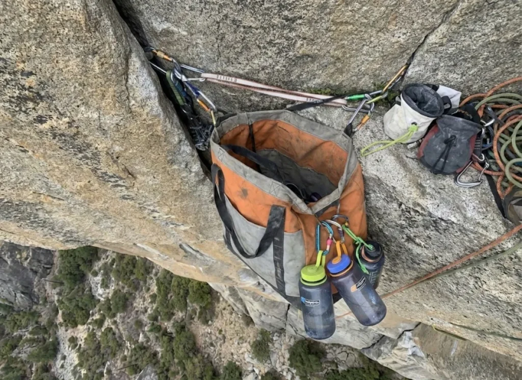A haul bag suspended on a cliff face with water bottles clipped to the outside loops for easy access.