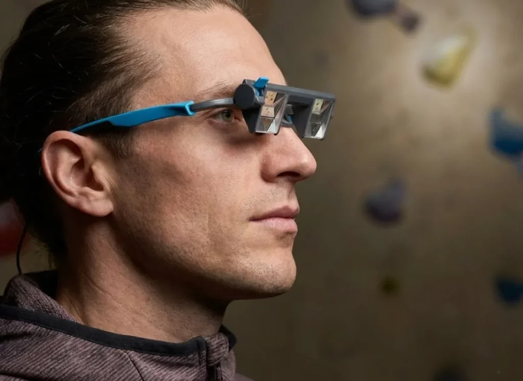 Profile view of a climber wearing PitchSix EyeSend glasses, with the climbing wall clearly reflected in the adjustable prism mechanism.