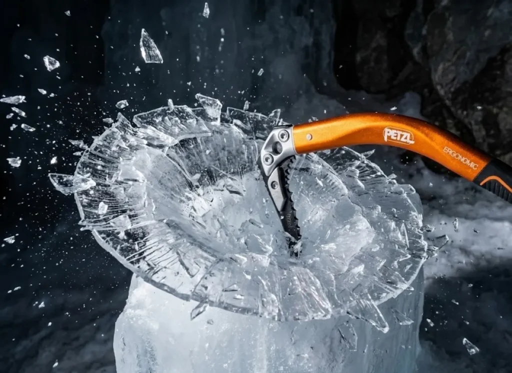 Close-up high-speed action shot of an ice axe striking brittle ice, causing a dinner-plate fracture with ice shards exploding outward, illuminated by dramatic strobe lighting.