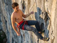 Adjust Your Leg Loops For Hanging Comfort A professional climber hanging comfortably at a vertical belay, inspecting the fit of their red Arc'teryx harness against a limestone cliff background.