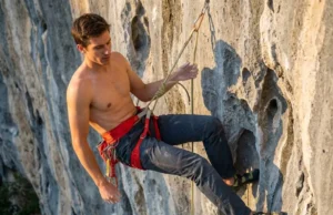 Adjust Your Leg Loops For Hanging Comfort A professional climber hanging comfortably at a vertical belay, inspecting the fit of their red Arc'teryx harness against a limestone cliff background.