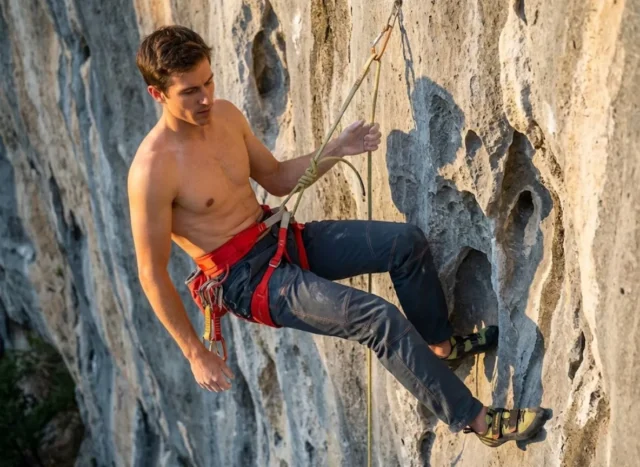 adjusting-leg-loops-hanging-comfort-cover A professional climber hanging comfortably at a vertical belay, inspecting the fit of their red Arc'teryx harness against a limestone cliff background.