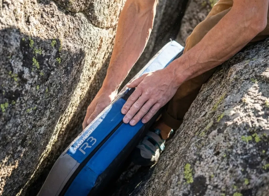 Close-up of a climber's hands using a Mad Rock R3 baffle pad to fill a deep hole in the talus field, demonstrating the Tetris method of leveling a landing zone.