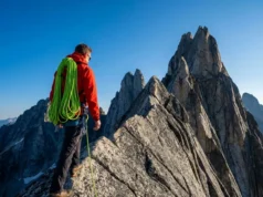 Butterfly Coils Carry Better For Long Approaches A professional climber on a granite ridge wearing a red Arc'teryx jacket and carrying a neon green climbing rope coiled like a backpack, illuminated by sharp morning sunlight.