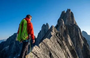 Butterfly Coils Carry Better For Long Approaches A professional climber on a granite ridge wearing a red Arc'teryx jacket and carrying a neon green climbing rope coiled like a backpack, illuminated by sharp morning sunlight.