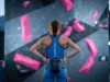 Read Competition Boulders Like A Route Setter A professional climber standing on competition mats analyzing a complex wall of pink volumes and dual-texture holds under dramatic stadium lighting.