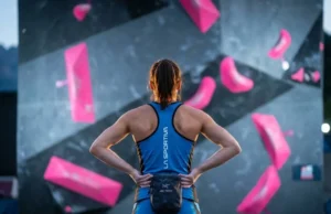 Read Competition Boulders Like A Route Setter A professional climber standing on competition mats analyzing a complex wall of pink volumes and dual-texture holds under dramatic stadium lighting.