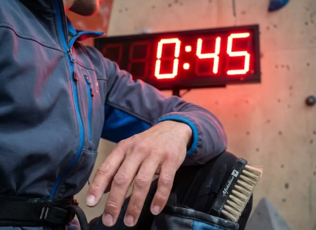 A climber resting on the mats during a competition rotation, looking at a digital timer counting down the final seconds of the round.