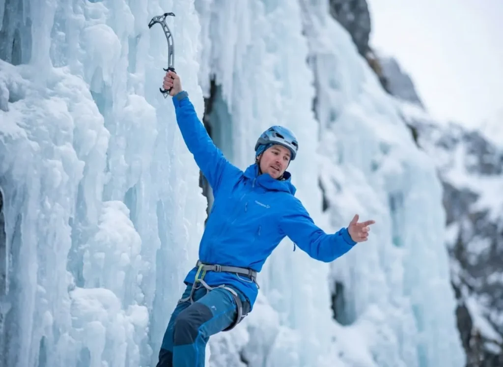 Ice climber resting on a vertical wall using a straight-arm skeletal hang technique to prevent fatigue, wearing a blue Norrøna jacket.
