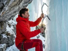 Swing Your Ice Tools With Less Energy Waste Elite ice climber in a red Arc'teryx jacket scaling a vertical blue ice pillar with Petzl Nomic tools, illuminated by dramatic alpine sunlight.