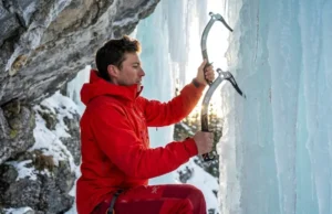 Swing Your Ice Tools With Less Energy Waste Elite ice climber in a red Arc'teryx jacket scaling a vertical blue ice pillar with Petzl Nomic tools, illuminated by dramatic alpine sunlight.