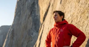 Why Free Soloing Is Not Just Climbing Without Ropes A professional climber in a red Arc'teryx jacket looking up at the base of El Capitan in Yosemite, illuminated by golden hour sunlight.