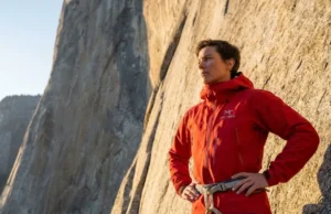Why Free Soloing Is Not Just Climbing Without Ropes A professional climber in a red Arc'teryx jacket looking up at the base of El Capitan in Yosemite, illuminated by golden hour sunlight.