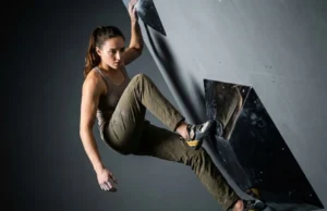 Grip Dual-Texture Holds Without Slipping Off A professional climber wearing Arc'teryx gear and La Sportiva shoes carefully placing a foot on a glossy black dual-texture bouldering volume in a dimly lit climbing gym.
