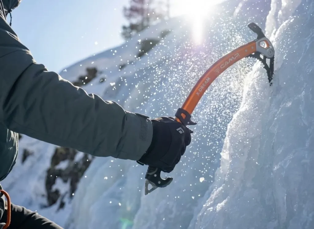 Action shot of an ice climber's gloved hand executing a wrist flick with a C.A.M.P. X-Dream tool, shattering ice chips in frozen motion.