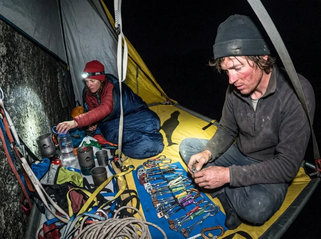 Climbers settling onto a portaledge bivy at night high on El Capitan, lit by a headlamp and camera flash