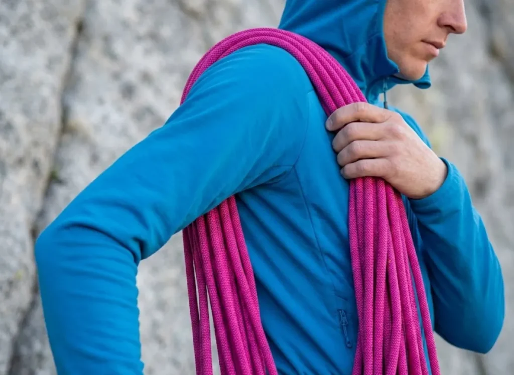 Side profile of a climber securing a pink climbing rope around their shoulders in a backpack configuration over a blue softshell jacket.