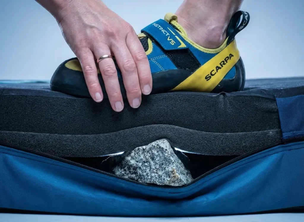 Close-up macro photography showing a climbing shoe compressing a crash pad to its limit, illustrating the danger of bottoming out on sharp rocks without adequate foam layering.