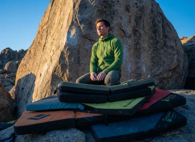 stack-pads-eliminate-gaps-landing-zones A professional climber stands confidently on a meticulously stacked system of Organic Climbing and Black Diamond crash pads at the base of a highball boulder during sunset in the Buttermilks.