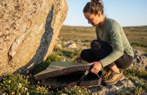 Where You Drop Your Pad Matters More Than You Think Female climber carefully placing Organic Climbing crash pad at base of granite boulder in alpine meadow