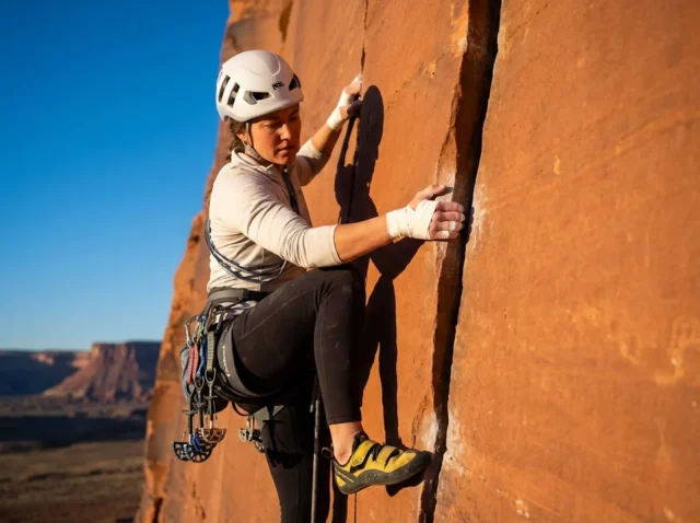 reading-crack-sizes-hand-jam-hero Climber placing a hand jam in a sandstone splitter crack at Indian Creek with tape gloves and cams on harness