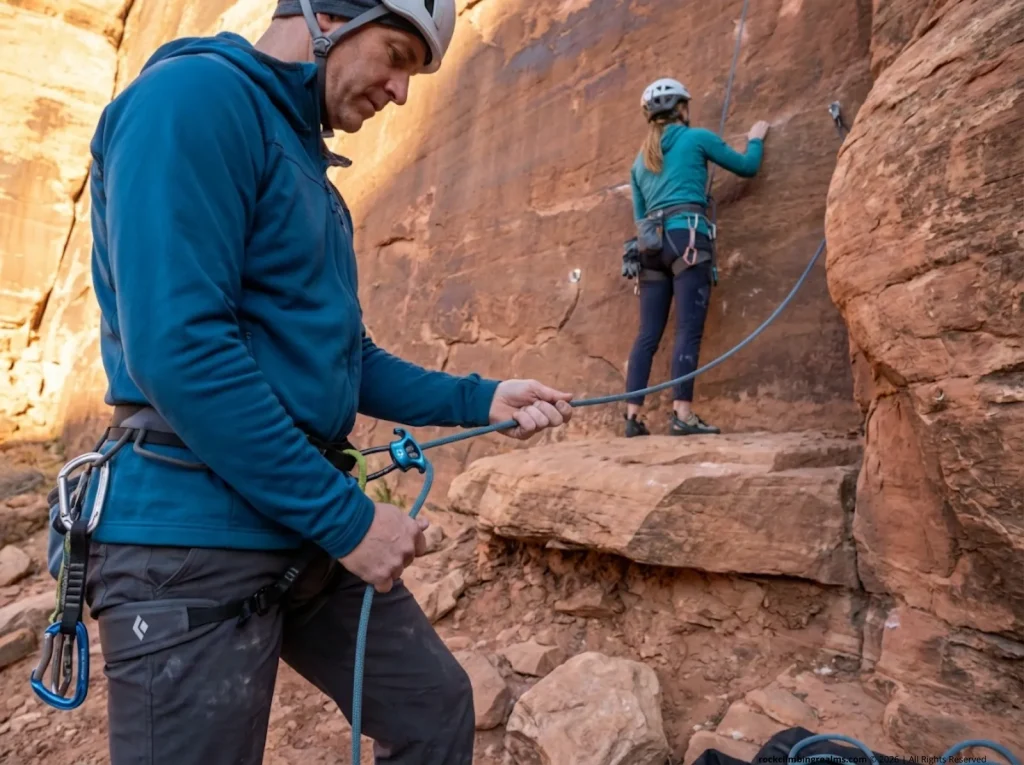 Belayer maintaining tight stance with ATC at base of sport climb, climber low on wall near ground ledge 