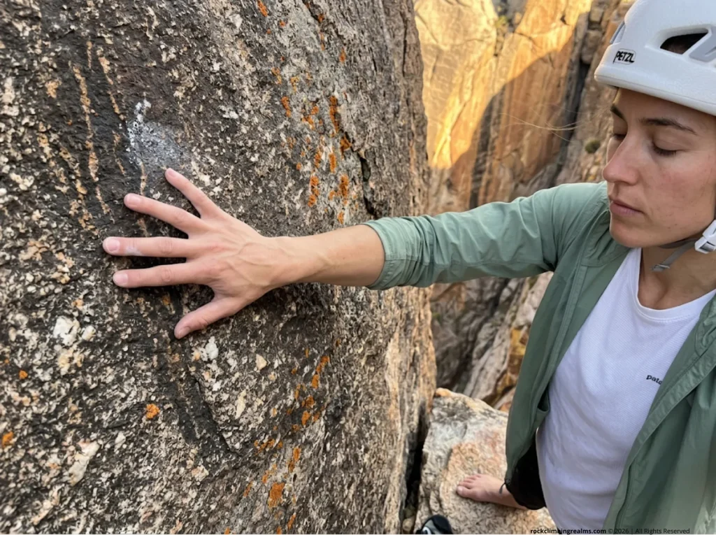 Climber demonstrates conduction heat loss by pressing bare palm against cold granite, understanding the physics of alpine thermal transfer firsthand