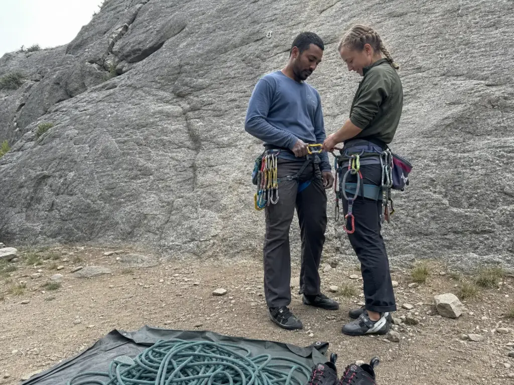 Two climbers running a gear check at the base of a route, checking carabiner orientation before climbing