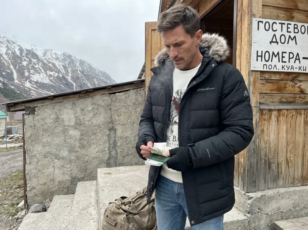 Climber in a down parka outside a Terskol guesthouse handling cash and passport for an Elbrus climb