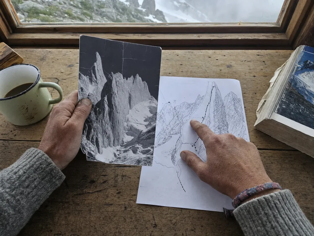 Climber comparing an old black-and-white summit photo against a route topo of Cerro Torre on a hut table