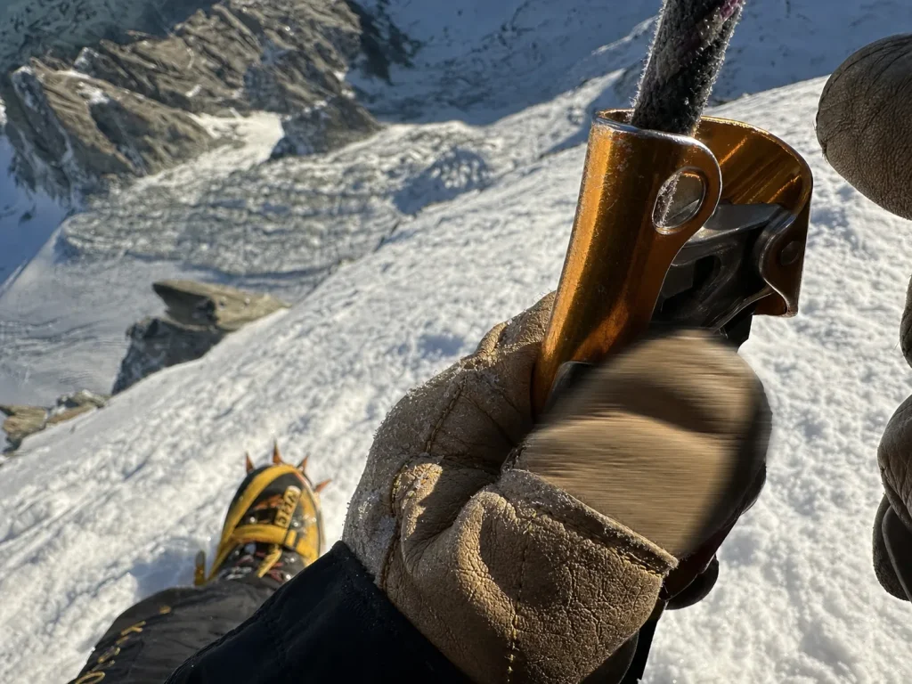 Gloved hands working a mechanical ascender on a fixed rope, crampons below