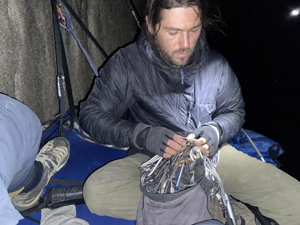Climbers organizing gear on a portaledge at dusk on El Capitan lit by harsh flash